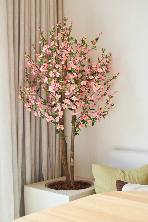 La plante de cerisier artificielle 120 cm rose, avec ses fleurs de cerisier roses, se trouve à côté d'une table en bois dans une pièce lumineuse et moderne, avec un rideau beige et un canapé clair derrière.