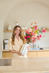 Een vrouw in een beige outfit staat in een moderne keuken. Ze glimlacht en houdt het boeket ‘Wild Peach’ van Zijden kunstbloemen vast, dat in een strakke witte vaas is geschikt en vol kleurrijke bloemen staat. Op de achtergrond zijn planken te zien met keukengerei en een koffiezetapparaat.