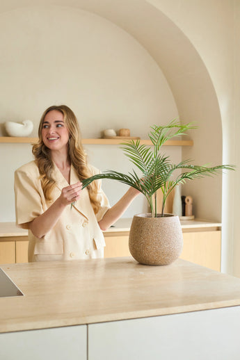 Une femme aux longs cheveux blonds sourit alors qu’elle s’occupe d’un palmier Chamaedorea artificiel en pot de 18 pouces dans une cuisine moderne aux tons neutres avec un décor minimaliste.