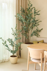 Deux plantes artificielles d'eucalyptus artificiel de 150 cm, placées à côté de rideaux transparents et de murs beiges, associées à une table à manger et des chaises en bois clair dans un intérieur minimaliste et moderne.