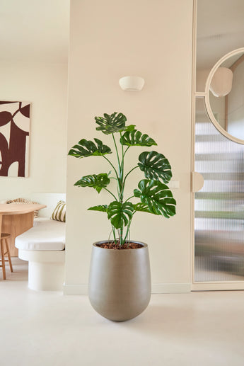 La plante artificielle Monstera de 120 cm est placée dans un espace de vie moderne et minimaliste avec des murs neutres, un miroir rond, des meubles en bois et des œuvres d'art murales abstraites.