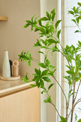 Une plante artificielle Pieris Japonica de 150 cm se dresse près d'une fenêtre à côté d'un meuble en bois clair contenant des bouteilles en céramique et un moulin à poivre, ajoutant une touche minimaliste à la pièce lumineuse et aérée.