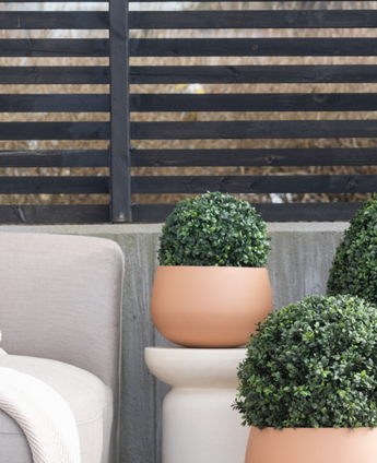 Trois topiaires rondes UV de style romain en buis artificiel de 30 cm se trouvent sur un patio près d'une chaise d'extérieur beige, contre une clôture moderne à lattes noires et des arbres nus.