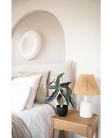 Een gezellige slaapkamer met neutrale tinten heeft een bed met wit linnen, een houten nachtkastje met de Kunst Begonia 48 cm in sierpot zwart en een geweven lamp, en ronde muurkunst met textuur boven het bed.