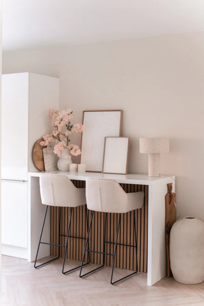 Een minimalistische thuisbar met twee beige krukken met kussens, een witte toonbank, ingelijste kunst, een tafellamp, neutraal getinte decoratie en Kunst Kersenbloesem 100 cm licht roze in vazen tegen een lichtgekleurde muur.