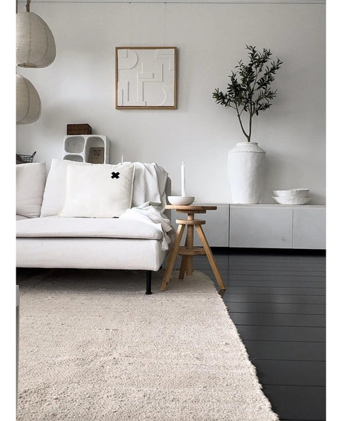 Un salon minimaliste avec un canapé de couleur claire, un tapis neutre, un buffet blanc, un olivier artificiel de 120 cm comme élément décoratif, une décoration murale abstraite et des tabourets en bois - le tout dans une douce ambiance méditerranéenne aux tons neutres.