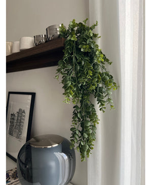 De Kunst hangplant Pannenkoek 61 cm hangt aan een houten plank boven een grijze glazen vaas, met kopjes en schalen ernaast. Een ingelijste zwart-wit stadsschets leunt tegen de muur bij een wit gordijn.
