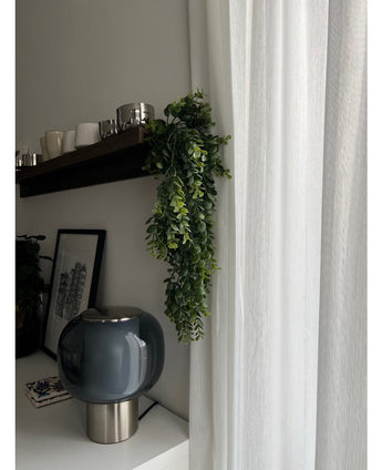 Een Kunst hangplant Pannenkoek 61 cm hangt aan een donkere houten plank boven een witte kast met een rookglazen lamp en een ingelijst zwart-wit stadsgezicht. Naast de plank hangt een wit gordijn.