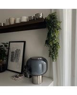 Een modern tafelblad met een lamp van donker glas, een ingelijste schets en onderzetters ligt onder een houten plank met kopjes en mokken. De Kunst hangplant Pannenkoek 61 cm hangt aan de plank, terwijl transparante gordijnen zacht natuurlijk licht binnenlaten.