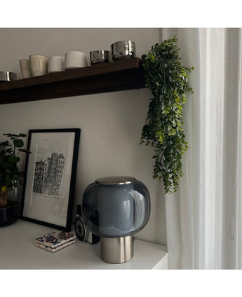 Een modern tafelblad met een lamp van donker glas, een ingelijste schets en onderzetters ligt onder een houten plank met kopjes en mokken. De Kunst hangplant Pannenkoek 61 cm hangt aan de plank, terwijl transparante gordijnen zacht natuurlijk licht binnenlaten.