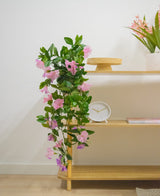 Een houten plank met Kunst hangplant Petunia 70 cm paars, een witte klok, een houten standaard, een boek en een vaas met roze bloemen tegen een witte muur.
