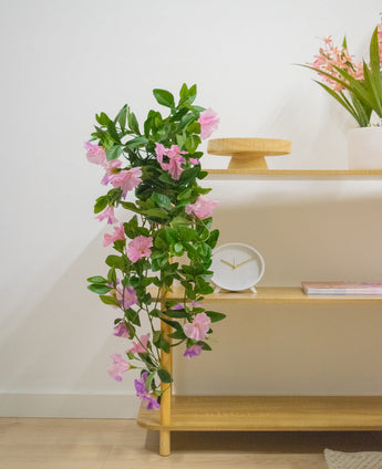 Een houten plank met Kunst hangplant Petunia 70 cm paars, een witte klok, een houten standaard, een boek en een vaas met roze bloemen tegen een witte muur.