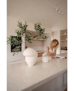 Een vrouw met krullend blond haar in een lichte outfit kijkt naar een open tijdschrift op een beige aanrecht. Op de voorgrond staat een sculpturale witte vaas met het Kunst olijfblad 90 cm UV, die een mediterrane flair geeft aan de minimalistische keuken.
