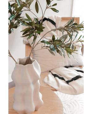Un vase sculpté blanc contient une feuille d'olivier Art 90 cm UV avec des feuilles vertes sur une table en bois clair. Derrière elle, une chaise beige et deux coussins de couleur crème aux rayures noires éclatantes rehaussent l'atmosphère méditerranéenne.
