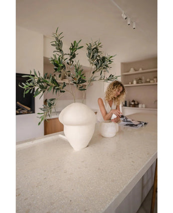 In een minimalistische keuken staat een grote crèmekleurige structuurvaas met een 90 cm groot brandvertragend kunst olijfblad op het aanrecht, terwijl een vrouw met krullend haar een tijdschrift leest bij open planken vol keramiek.