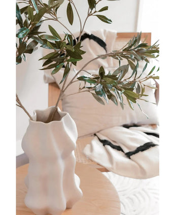 Een witte, sculpturale vaas met Kunst olijfblad 90 cm brandvertragend staat op een lichte houten tafel. Daarachter staan twee crèmekleurige kussens met zwarte strepen op een beige stoel, die knus comfort toevoegt aan deze moderne ruimte.