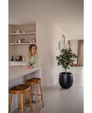 Een vrouw met krullend blond haar glimlacht achter een aanrecht met twee houten krukjes. De Kunst sinaasappelboom 120 cm in een pot voegt charme toe aan de moderne, minimalistische ruimte die is ingericht in neutrale tinten.