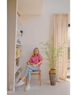 Een vrouw in een roze trui en spijkerbroek zit op een houten stoel naast de Kunst varen plant 140 cm, in de buurt van een boekenplank en een groot raam met beige gordijnen in een lichte, moderne woonruimte.