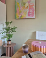 In een kamer met een pastelthema staan een Kunst-varenplant van 85 cm op een roze bijzettafeltje, een paars-gouden kopje, een roze schaal met geschulpte rand, een open notitieboekje en aan de muur een abstract schilderij met groene, roze en gele tinten.