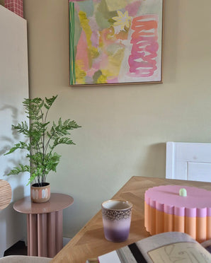 In een kamer met een pastelthema staan een Kunst-varenplant van 85 cm op een roze bijzettafeltje, een paars-gouden kopje, een roze schaal met geschulpte rand, een open notitieboekje en aan de muur een abstract schilderij met groene, roze en gele tinten.