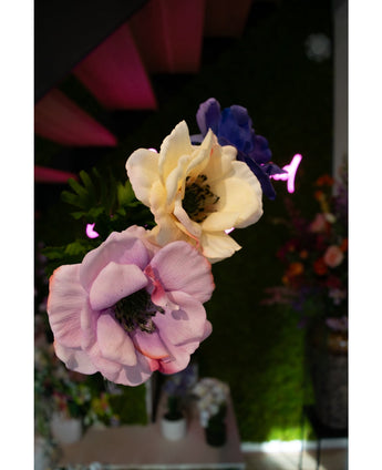 Close-up van de Kunstanemoon Real Touch paars 52cm kunstbloem, verticaal gerangschikt met andere bloemen, tegen een vage achtergrond van groen gebladerte, levendige bloemen en roze neonlichten.