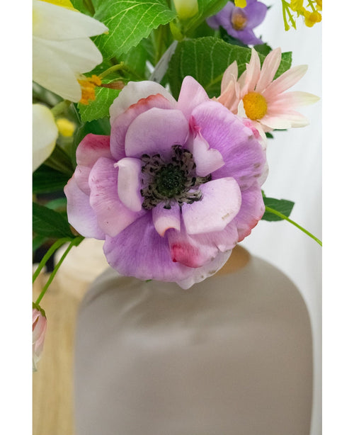 Gros plan de l'Anémone Artificielle Real Touch violet 52cm, une fleur violet clair et rose au cœur sombre, entourée de feuilles vertes et de fleurs aux couleurs pastel dans un vase blanc mat, sur un fond légèrement flou.