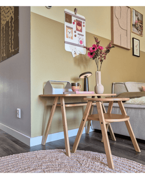 Een moderne woonkamerhoek met een lichte houten salontafel met daarop de Kunstbloem Anemoon vertakt 56 cm roze in een roze vaas, samen met een lamp en boeken. Ernaast staat een beige bank, geaccentueerd door muurkunst en een galerieachtig fotobord op de gele muur.