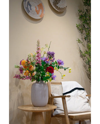 Un vase blanc avec cornouiller artificiel Real Touch Jaune Clair 74 cm se dresse sur une table d'appoint en bois à côté d'une chaise en bois clair avec un coussin blanc et un tapis aux bords noirs, accentués par une décoration murale tissée et une plante verte en fond.