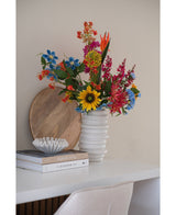 Un bouquet de fleurs d'œillets artificiels de 70 cm bleu dans un vase en céramique blanche se trouve sur un bureau blanc à côté de livres empilés et d'un petit bol décoratif, avec un plateau rond en bois appuyé contre le mur derrière lui.