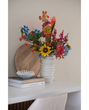 Un bouquet de fleurs d'œillets artificiels de 70 cm bleu dans un vase en céramique blanche se trouve sur un bureau blanc à côté de livres empilés et d'un petit bol décoratif, avec un plateau rond en bois appuyé contre le mur derrière lui.
