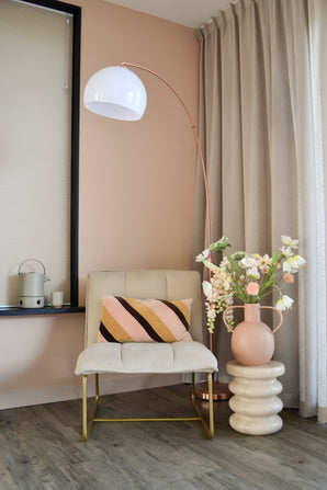 Een moderne, gezellige hoek met een crèmekleurige stoel met een gestreept kussen, een hoge boogvormige vloerlamp, beige gordijnen, een houten vloer en een ronde vaas met Kunstbloem Fritillary 94 cm wit bloemen op een gebeeldhouwde bijzettafel.