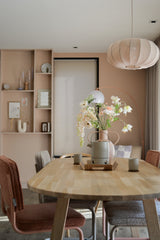 Une salle à manger moderne avec une table en bois clair, quatre chaises avec coussins, la fleur artificielle Fritillaire 94 cm blanche dans un vase, des tasses en céramique, des murs rose pastel, des étagères intégrées avec décor et une grande lampe ronde suspendue au-dessus de votre tête.