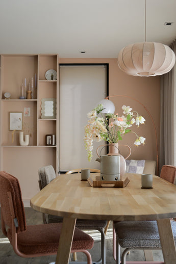 Une salle à manger moderne avec une table en bois clair, quatre chaises avec coussins, la fleur artificielle Fritillaire 94 cm blanche dans un vase, des tasses en céramique, des murs rose pastel, des étagères intégrées avec décor et une grande lampe ronde suspendue au-dessus de votre tête.
