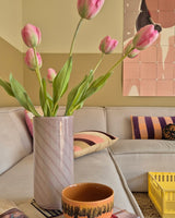 Een Kunstbloem Franse Tulp van 63 cm in roze staat in een vaas op de tafel naast een oranje kopje in een gezellige woonkamer met een crèmekleurige bank, kussens met patronen, een gele krat en abstracte wandkunst op de achtergrond.