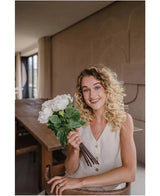 Een glimlachende vrouw met krullend blond haar in een mouwloos beige topje zit in een moderne kamer en houdt een Zijden kunstbloemen boeket Peony Wit vast. Ze kijkt naar de camera met een houten tafel zichtbaar op de achtergrond.