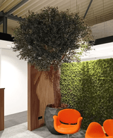 De Kunstboom Olijf 300 cm staat binnen naast een houten paneel en groene moswand, met twee oranje moderne stoelen ervoor onder een industrieel plafond.