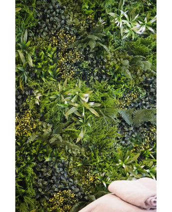 Een weelderige, gestructureerde achtergrond wordt gecreëerd door de Kunsthaag Freshness 100x100 cm UV verticale tuinmuur, dichtbegroeid met groen gebladerte. Rechtsonder zijn twee gevouwen beige handdoeken zichtbaar.