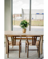 Een houten tafel met twee bijpassende stoelen staat op een rond kleed bij grote ramen. Een Kunstplant Baby Schefflera UV 45 cm siert de tafel, met uitzicht op gebouwen en een hek buiten.