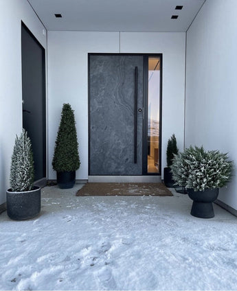Une porte d'entrée sombre et moderne est encadrée par des murs blancs et flanquée d'une pyramide de buis artificiel UV de 90 cm, qui offre une protection UV. Une légère neige recouvre le sol, tandis que ces plantes de jardin élégantes créent une entrée soignée et minimaliste.