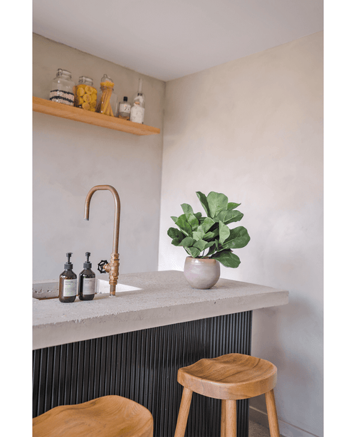 Un plan de travail moderne avec un robinet en laiton, deux distributeurs de savon, la plante en pot Artificielle Feuille de Tabac UV 45 cm et deux tabourets en bois. Une étagère flottante avec des pots et des bouteilles est montée sur un mur de couleur claire.