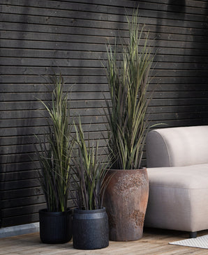Trois plantes artificielles de gazon artificiel Bordeaux UV 150 cm de différentes hauteurs se trouvent à côté d'un canapé de couleur claire sur un parquet en bois, avec un mur en bois sombre en arrière-plan.