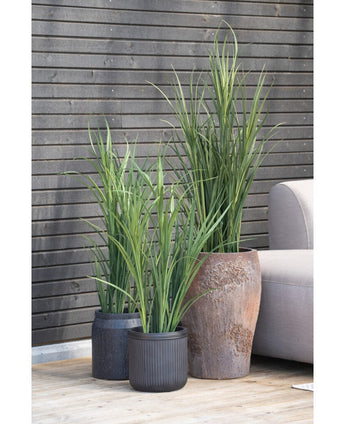 Une plante artificielle de 90 cm de gazon artificiel vert UV dans un pot noir se dresse sur un plancher en bois à côté d'un canapé d'extérieur gris, avec un mur à lattes de bois sombre en arrière-plan.