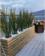 Een houten plantenbak met Kunstgras Groen UV 90cm en witte stenen staat op een houten vlonder naast een zwarte plantenbak met kleurrijke hortensia's, met glazen balustrades en uitzicht op de bergen op de achtergrond.