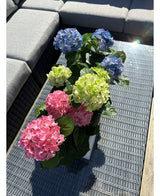 De Kunstplant Hortensia Groen UV 35 cm staat op een rieten tafel met glazen tafelblad naast een grijze buitenbank, met realistische groene hortensia's die oplichten in het zonlicht.