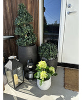 Een Kunstplant Hortensia Groen UV 35 cm in een witte pot met geelgroene bloemen staat op de veranda bij een bruine deur, naast twee planten in een zwarte pot en twee decoratieve lantaarns met kaarsen.