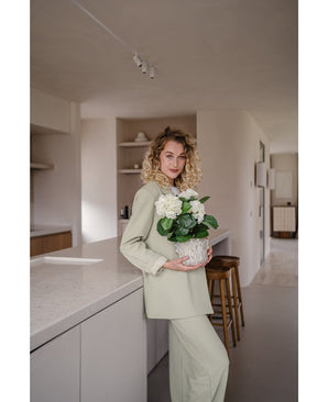 Une femme aux cheveux blonds bouclés, vêtue d'une tenue vert clair, se tient dans une cuisine moderne avec dans ses mains une plante d'hortensia artificiel blanc UV de 35 cm – avec des fleurs blanc crème – et sourit à la caméra.