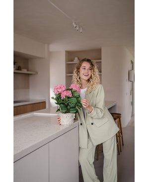 Een vrouw in een lichtgroen pak staat in een moderne keuken en glimlacht terwijl ze de Kunstplant Hortensia Roze UV 35 cm op het aanrecht vasthoudt.