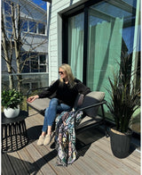 Een vrouw met zonnebril, zwart topje en blauwe jeans ontspant op een kussensstoel op een zonnig balkon met een drankje in de hand. De ruimte is voorzien van de Kunstplant Laurier UV d33 cm, een levendig decor, een kleurrijke deken en een kleine tafel met planten.