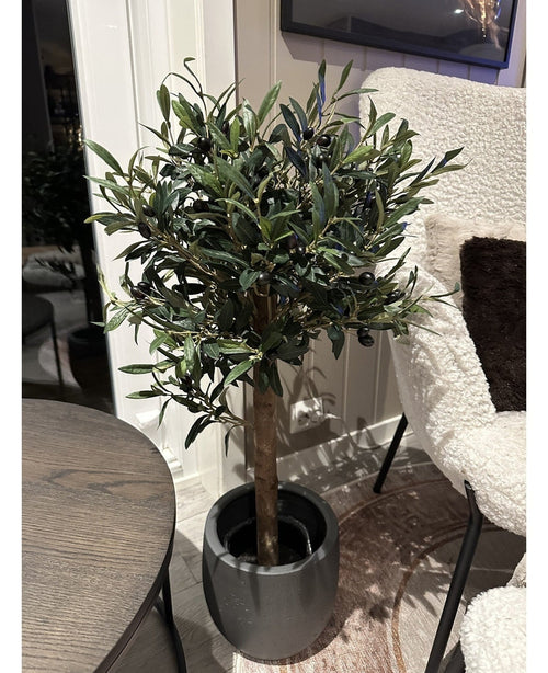 L'Olivier artificiel UV 85 cm, une plante artificielle compacte et facile d'entretien aux feuilles vert foncé et aux olives noires, se dresse en intérieur à côté d'une table ronde en bois et d'un fauteuil à la texture blanche.