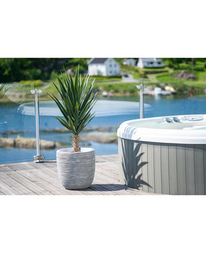 Een Kunstplant Yucca 100 cm UV staat op een houten dek naast een hot tub met glazen reling, met uitzicht op een rustig meer en groene heuvels bezaaid met huizen.
